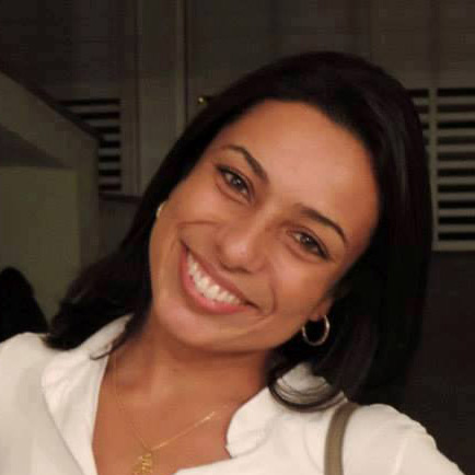 Foto do rosto de uma mulher, cabelos pretos de tamanho médio, tem a cabeça levemente inclinada para o lado direito e sorri abertamente. Usa brincos de argola e uma camisa de gola branca.